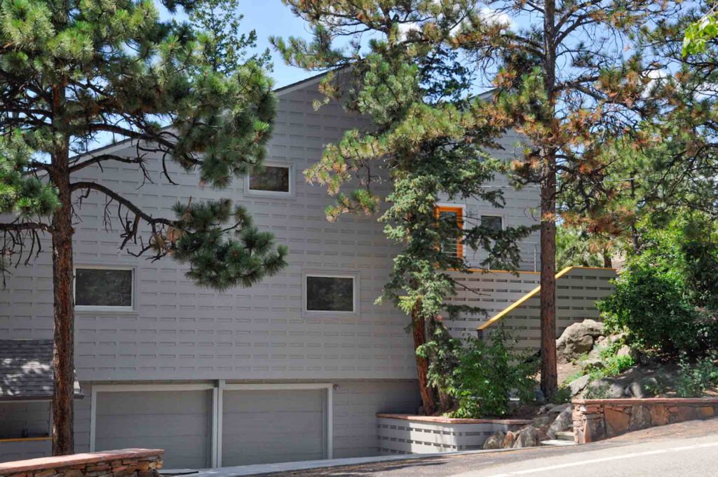 Two-story gray brick house with double garage, surrounded by pine trees and stone landscaping.