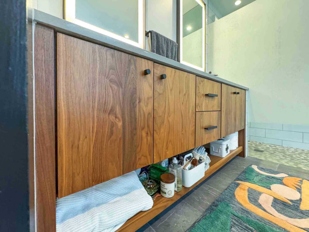 Wooden bathroom vanity with open shelf of towels and products, illuminated mirrors, and colorful rug.