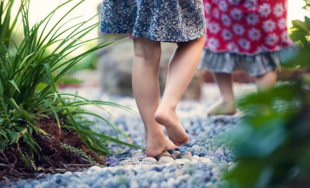 Two children in dresses walking barefoot on a pebble path with surrounding greenery.
