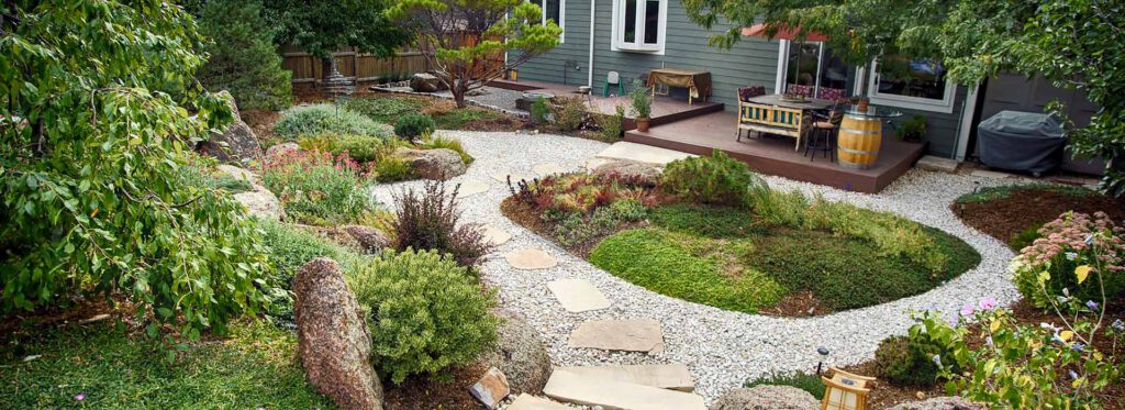 Backyard garden with gravel path, wooden deck, and outdoor seating Backyard garden with gravel path, stone steps, green plants, wooden deck, outdoor seating, and barbecue grill.
