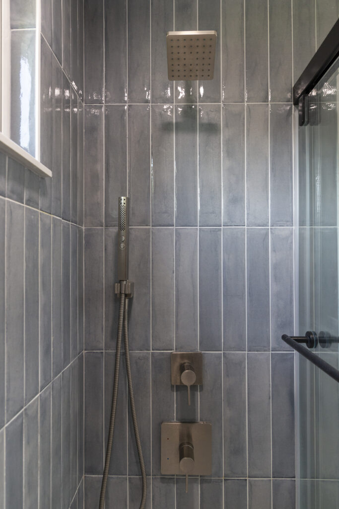 Shower area with vertical blue-gray tiles, square rain showerhead, handheld shower, controls, and glass door.