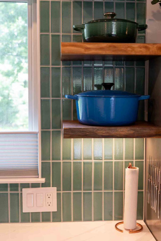 Kitchen corner with green tiles and wooden shelves Kitchen wall with green vertical tiles, wooden shelves holding green and blue pots, window, and paper towel holder.