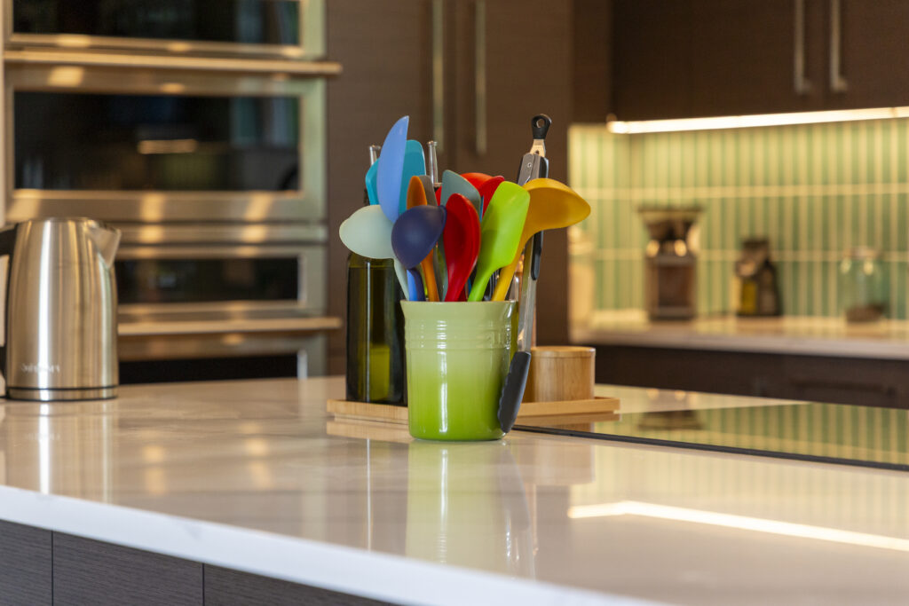 Kitchen countertop with colorful utensils in a green holder, electric kettle, and built-in ovens.