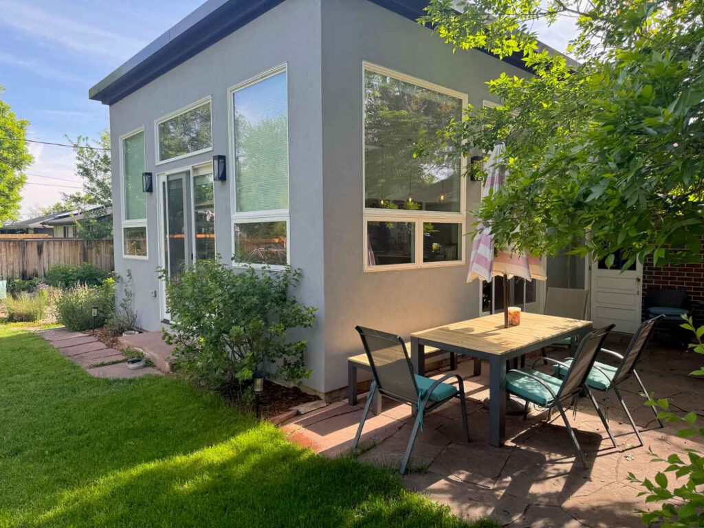 Gray modern building with large windows next to a patio with table, chairs, and striped umbrella.