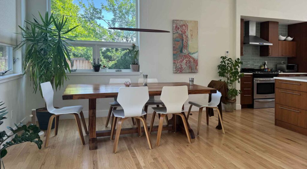 Wooden dining table with six white chairs in a bright kitchen area Dining area with wooden table, six white chairs, plants, large windows, and kitchen background.