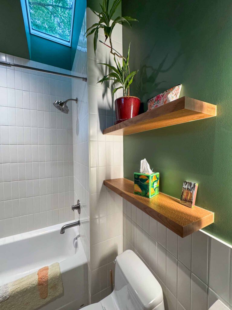 Bathroom with skylight, bathtub, and wooden shelves Bathroom with white tiled walls, bathtub, showerhead, skylight, wooden shelves, and green wall.