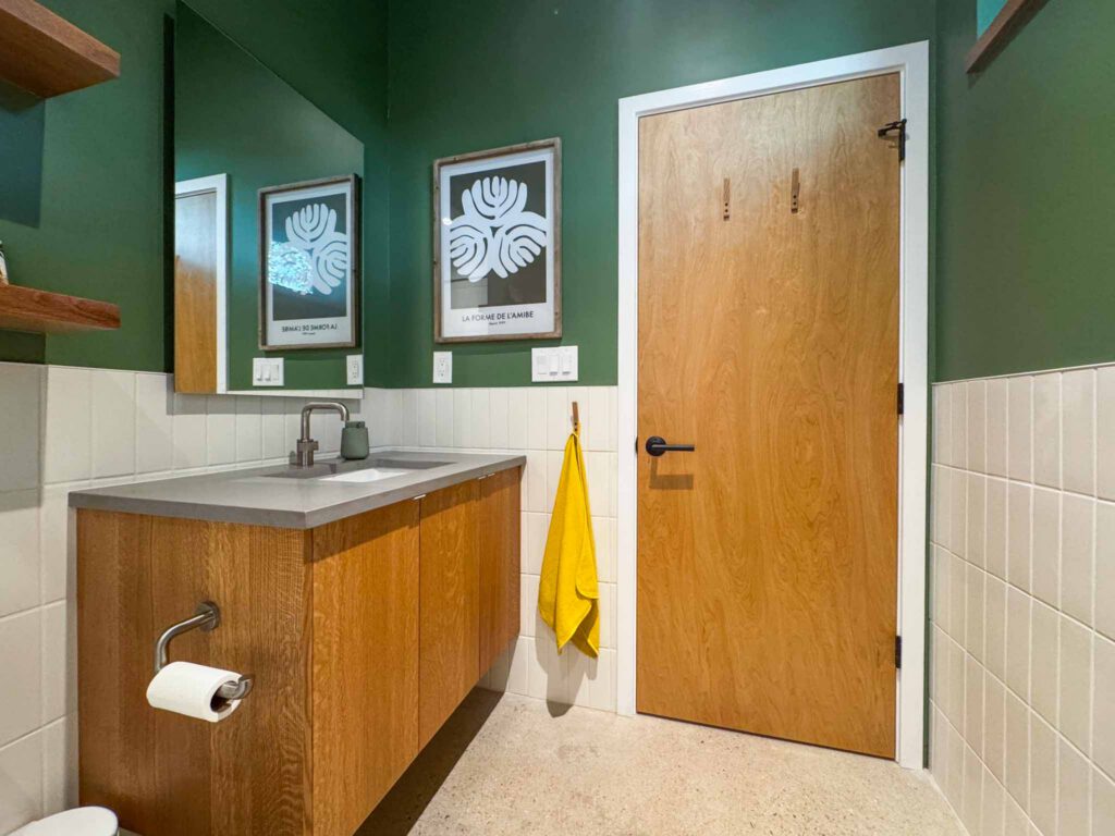 Modern bathroom with green walls and wooden vanity Modern bathroom with green upper walls, white tiles, wooden vanity, yellow towel, and framed artwork.