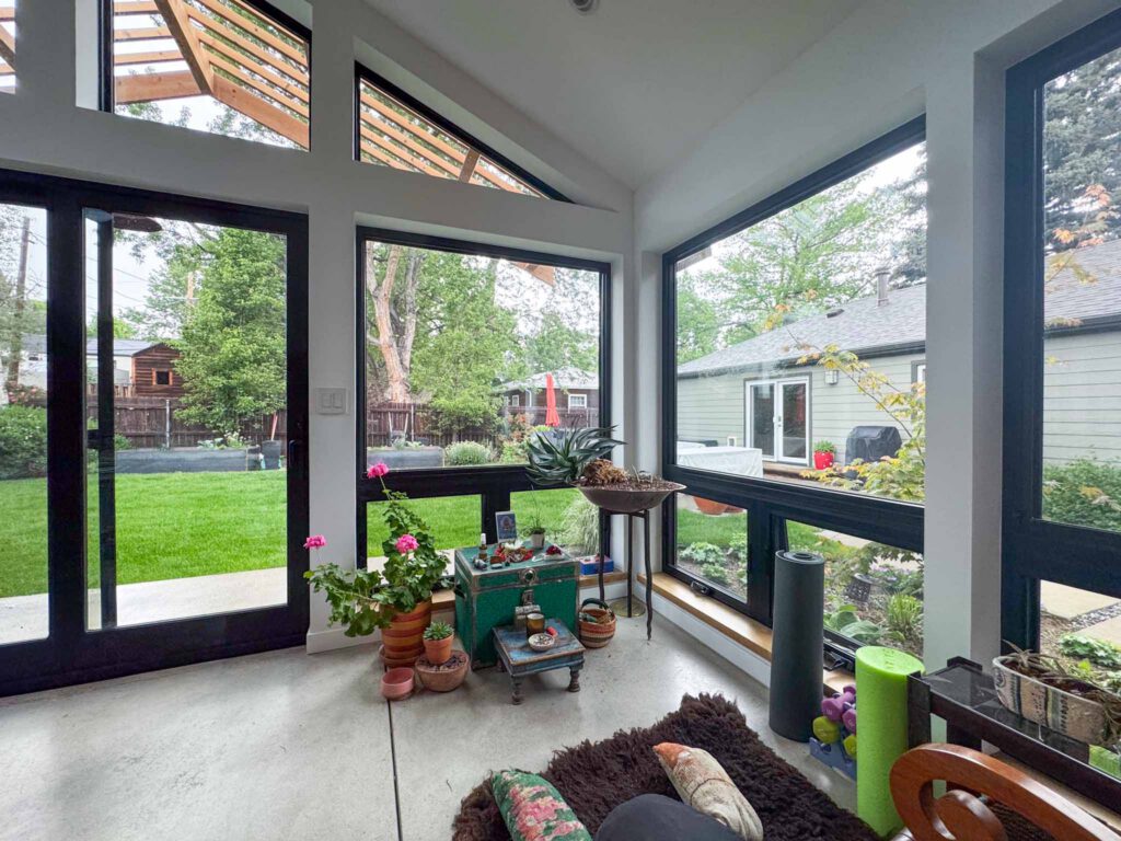 Sunlit room with plants and fitness equipment overlooking backyard patio Room with large black-framed windows, indoor plants, fitness gear, and view of backyard patio.