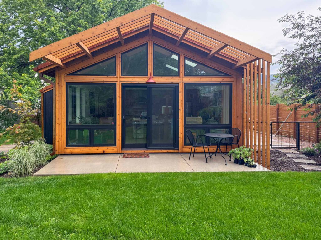 Modern wooden cabin with glass windows and patio seating Modern wooden cabin with large windows, concrete patio, black table, two chairs, and green lawn.
