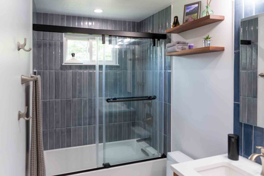 Modern minimalist bathroom with bathtub and sliding glass shower door Modern bathroom with bathtub, sliding glass shower door, gray tiled walls, and floating wooden shelves.