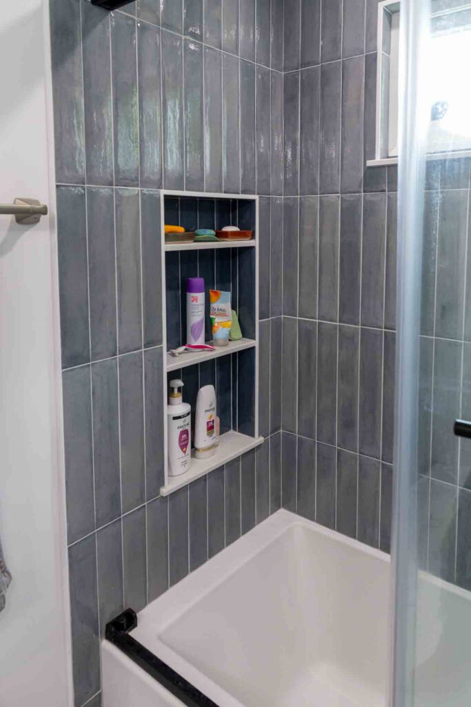 Shower with gray tiles, built-in shelves holding toiletries, white tub, and glass door.