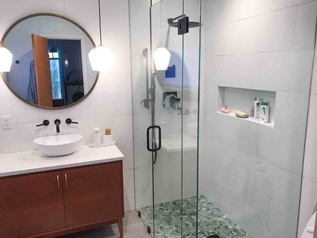 Modern bathroom with wooden vanity and glass shower enclosure Modern bathroom with wooden vanity, vessel sink, round mirror, pendant lights, and glass shower with tiled walls.
