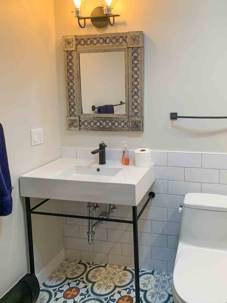 Small bathroom corner with patterned tiles and decorative mirror Bathroom corner with white sink, black faucet, patterned floor tiles, toilet, and a decorative mirror.
