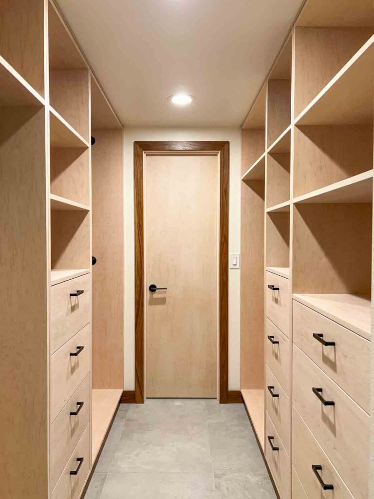 Narrow walk-in closet with light wood shelves, drawers with black handles, gray tile floor, and wooden door.