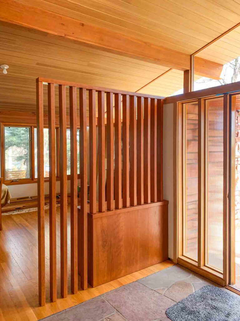 Wooden room interior with vertical slat partition, wooden floor, large windows, and glass door.