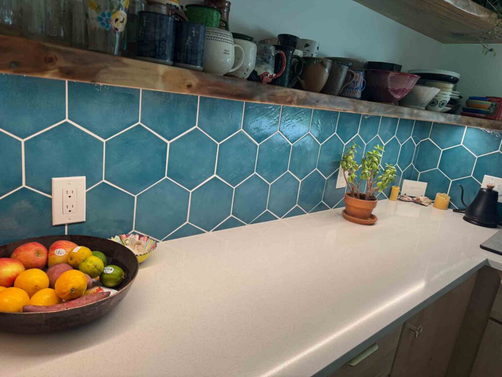 Kitchen countertop with teal hexagonal tile backsplash, wooden shelves, mugs, bowls, fruit bowl, plant, and kettle.