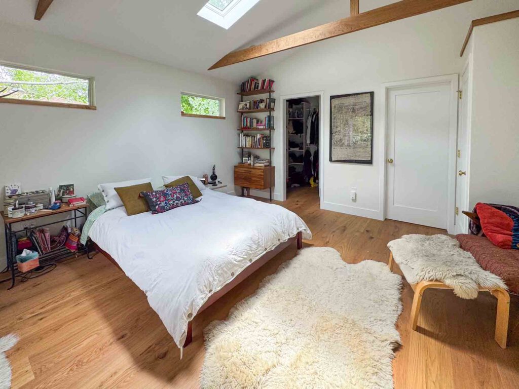 Minimalist bedroom with skylight and wooden flooring Bedroom with wooden floor, white bed and pillows, skylight, windows, shelving, and closet.