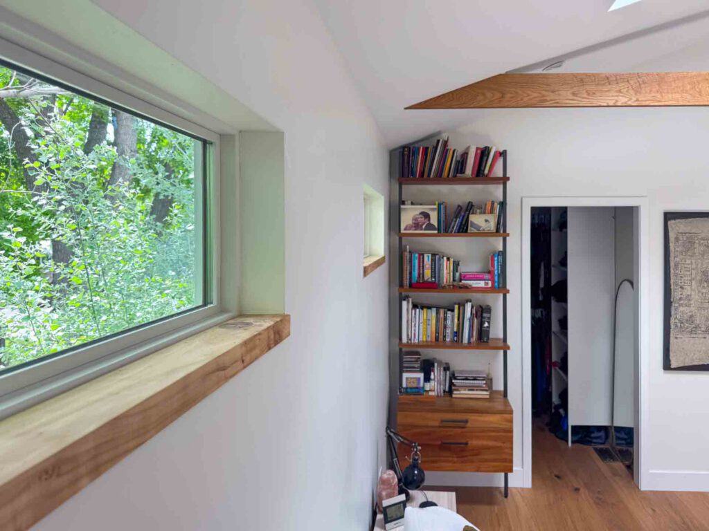 Minimalist room with bookshelf and large window Room with white walls, wooden floor, large window with trees outside, bookshelf, and doorway.