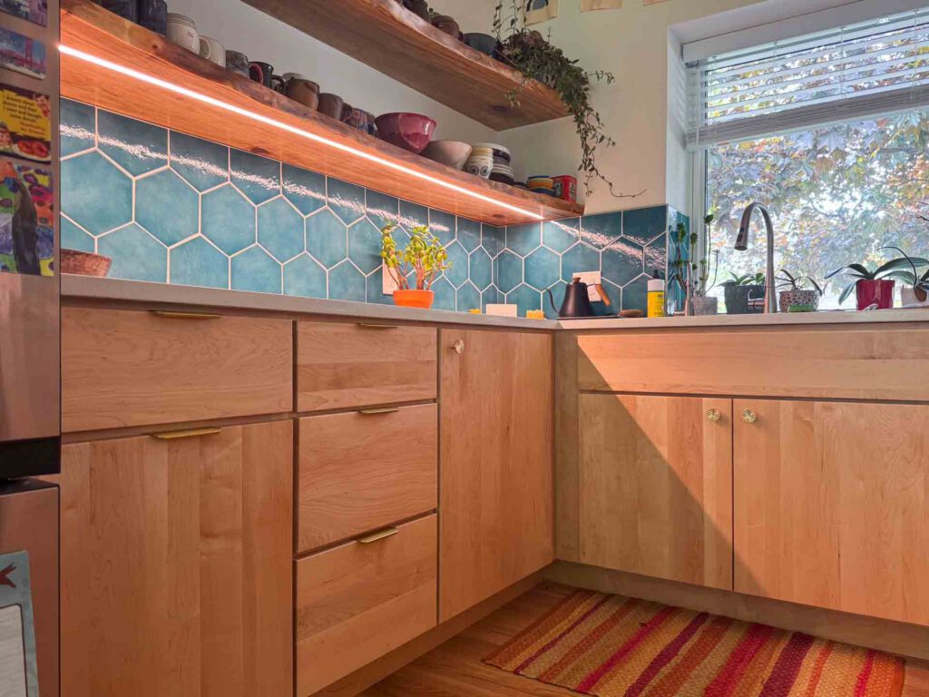 Kitchen corner with blue hex tile backsplash and wooden shelves Kitchen corner with light wood cabinets, blue hexagonal tile backsplash, and window above sink.
