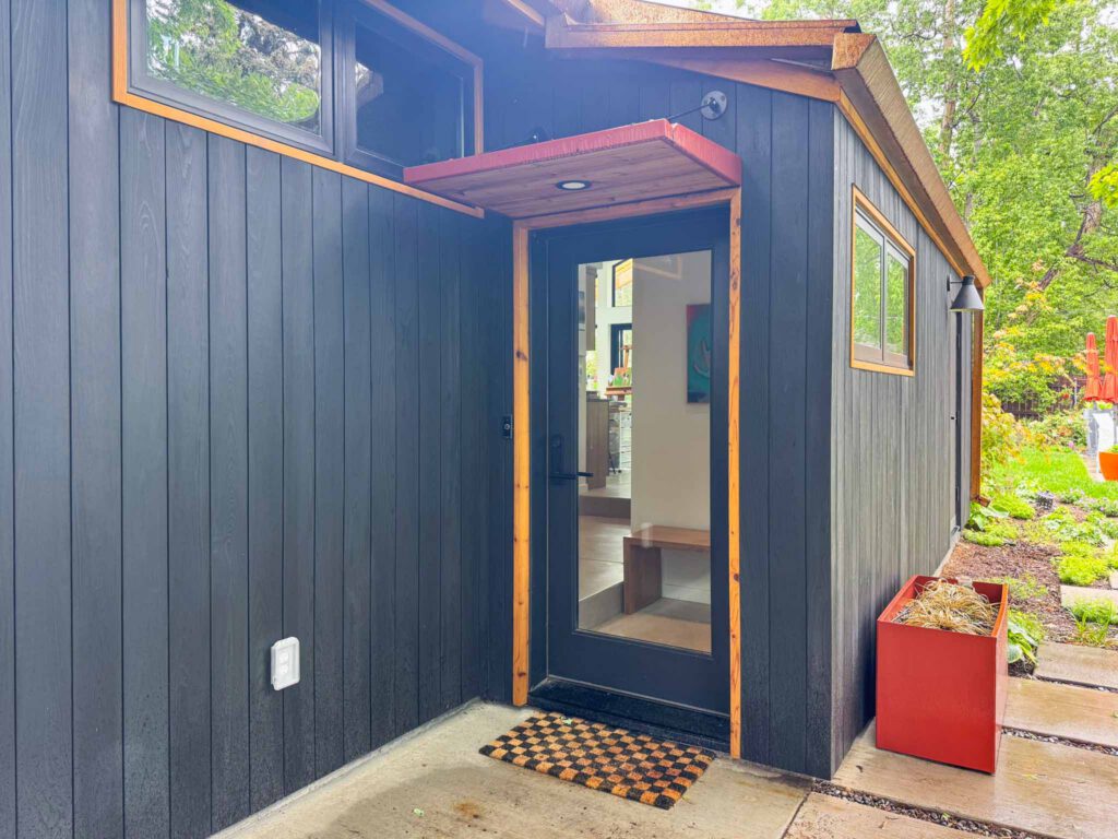 Modern building entrance with dark wood siding and glass door Modern black wood exterior entrance with glass door, wooden trim, checkered doormat, and red planter.
