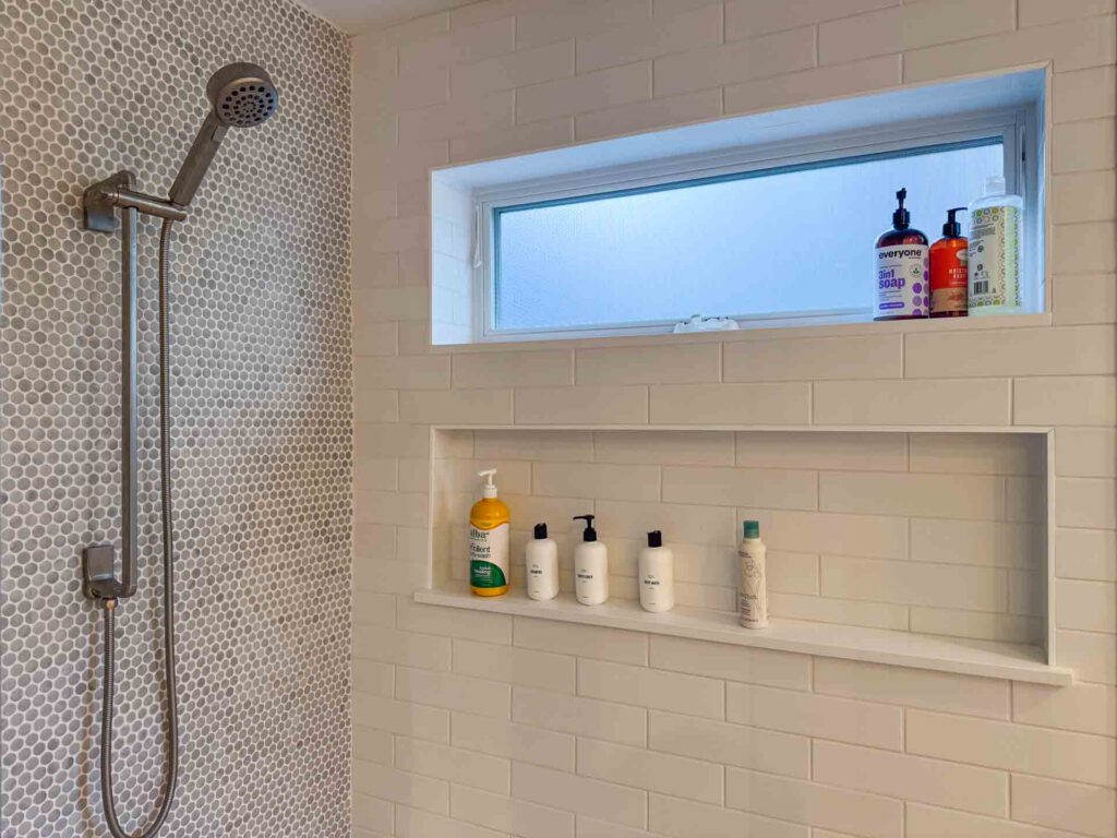 Modern shower area with tiled walls and recessed shelf Shower with wall-mounted showerhead, circular and rectangular tiles, recessed shelf, and frosted window with toiletries.