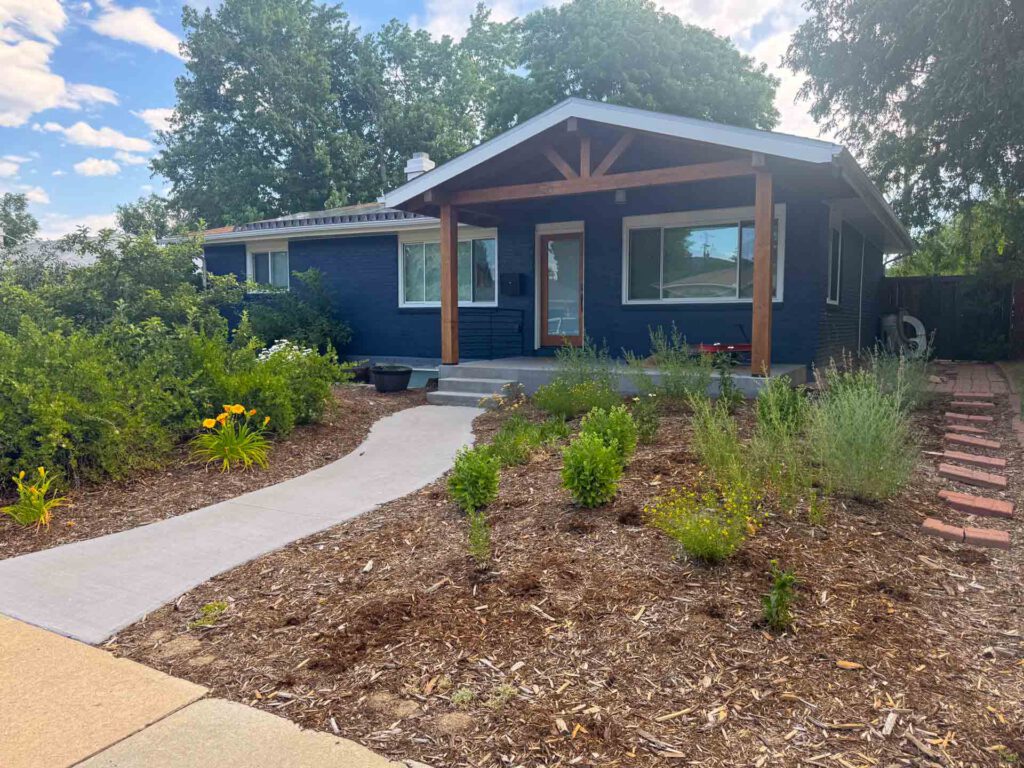 Single-Story Blue House with Covered Porch and Landscaped Yard Single-story dark blue house with wooden porch, concrete walkway, mulch, plants, and brick path.
