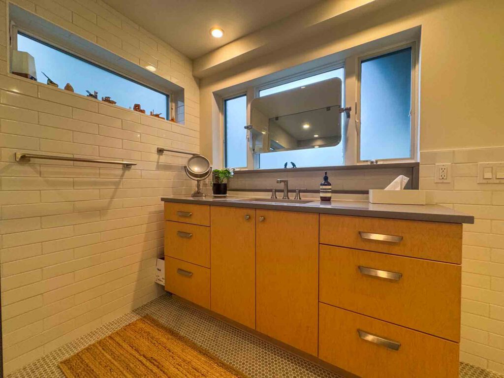 Modern bathroom vanity with light wood cabinets and gray countertop Bathroom vanity with wood cabinets, gray countertop, sink, mirror, white tile walls, and small windows.